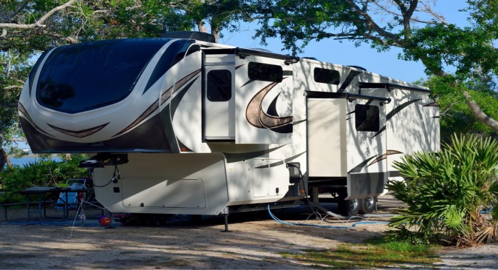 Large fifth-wheel RV with slide-outs parked at a shaded campsite beneath oak and palm trees